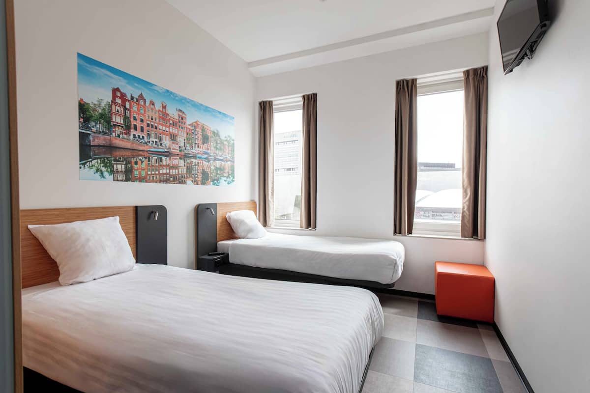 Une chambre d’hôtel avec deux lits, une télévision, un tableau représentant un paysage urbain et une fenêtre avec des rideaux.