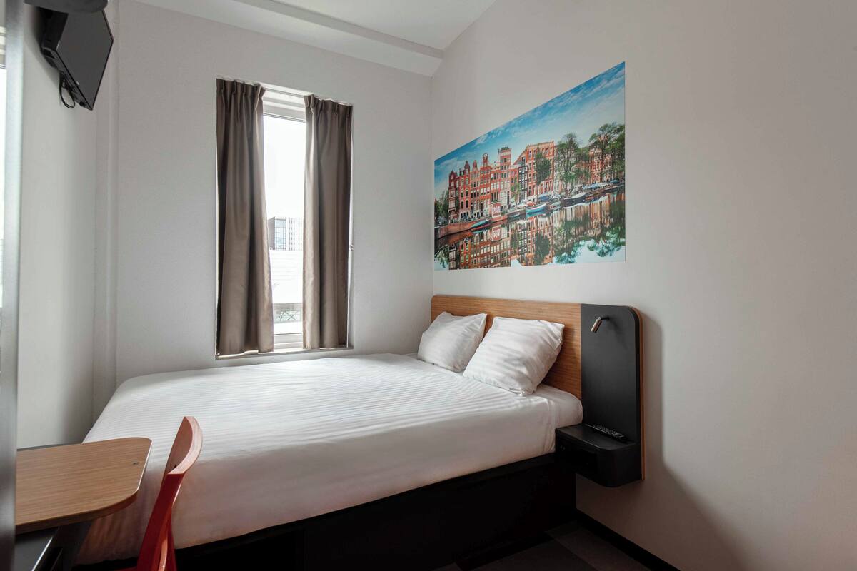 Une chambre d’hôtel avec un lit, un bureau, une chaise, un tableau représentant un paysage urbain et une fenêtre avec des rideaux.