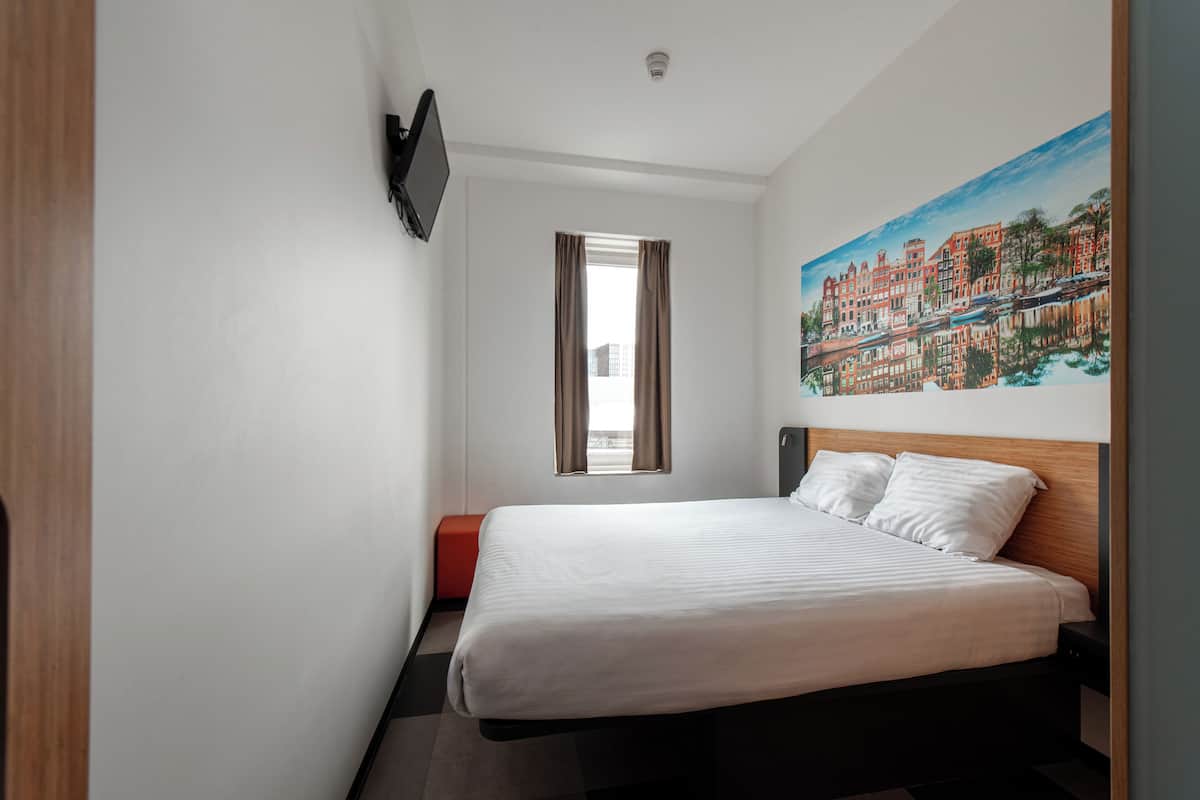 Une chambre d’hôtel avec un lit, une fenêtre avec des rideaux, un tableau accroché au mur et un téléviseur fixé au mur.
