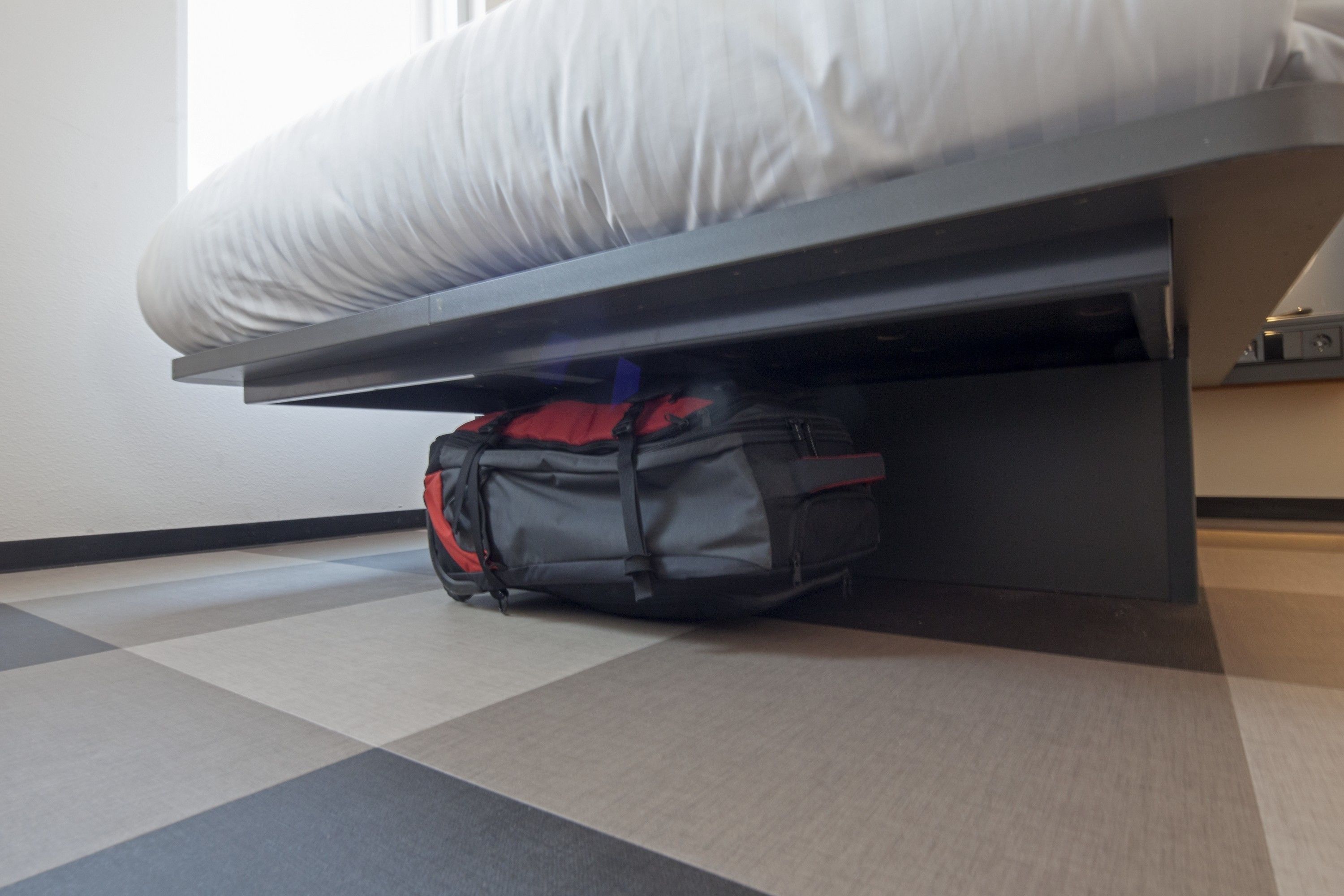 Una cama con colchón y estructura metálica negra. Una mochila roja y negra está debajo de la cama. El suelo tiene un estampado de cuadros.