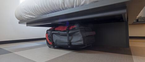 Una cama con colchón y estructura metálica negra. Una mochila roja y negra está debajo de la cama. El suelo tiene un estampado de cuadros.