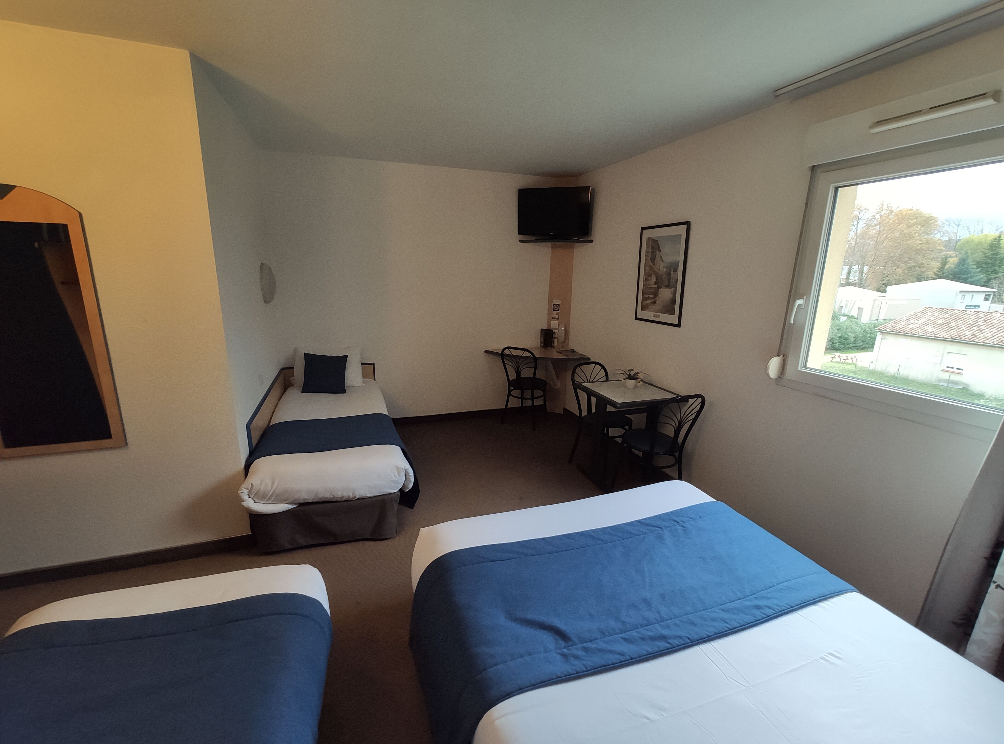 A hotel room with two single beds, a desk with a chair, a television mounted on the wall, and a window with a view of the outside.