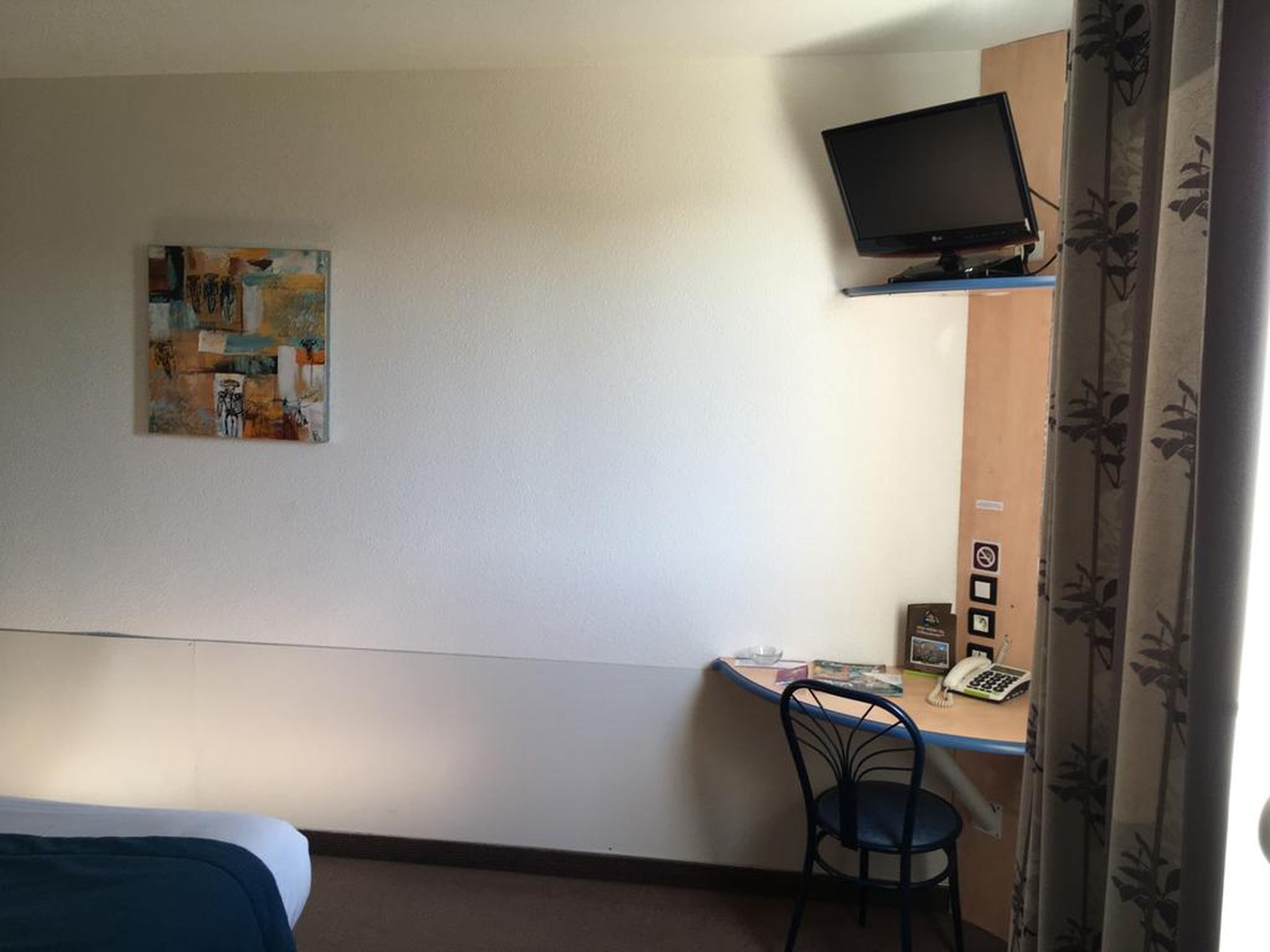 A hotel room with a bed, a desk with a chair, a television mounted on the wall, and a painting on the wall.