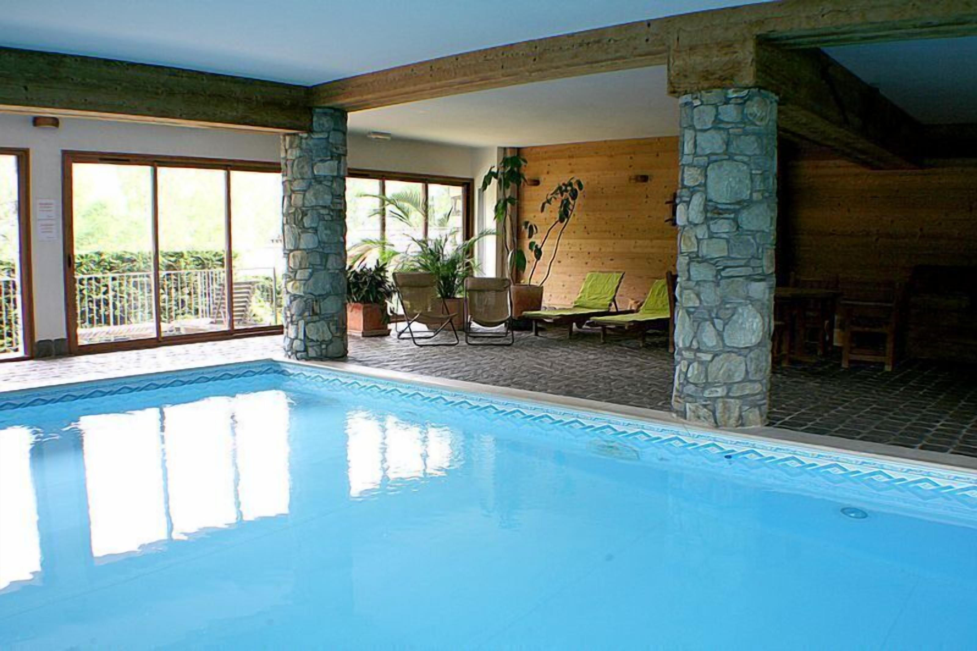 Une piscine intérieure agrémentée de piliers en pierre, un espace détente avec des fauteuils et une table, et de grandes fenêtres laissant entrer la lumière naturelle.