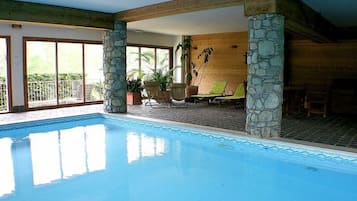 Une piscine intérieure agrémentée de piliers en pierre, un espace détente avec des fauteuils et une table, et de grandes fenêtres laissant entrer la lumière naturelle.