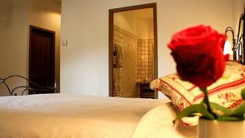 Un dormitorio con una cama, una mesita de noche, un florero con una rosa roja y un baño visible a través de una puerta abierta.