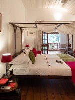 Uma cama de dossel com lençóis brancos e detalhes vermelhos, uma mesa de cabeceira com abajur, e vista para o exterior através de uma grande janela.