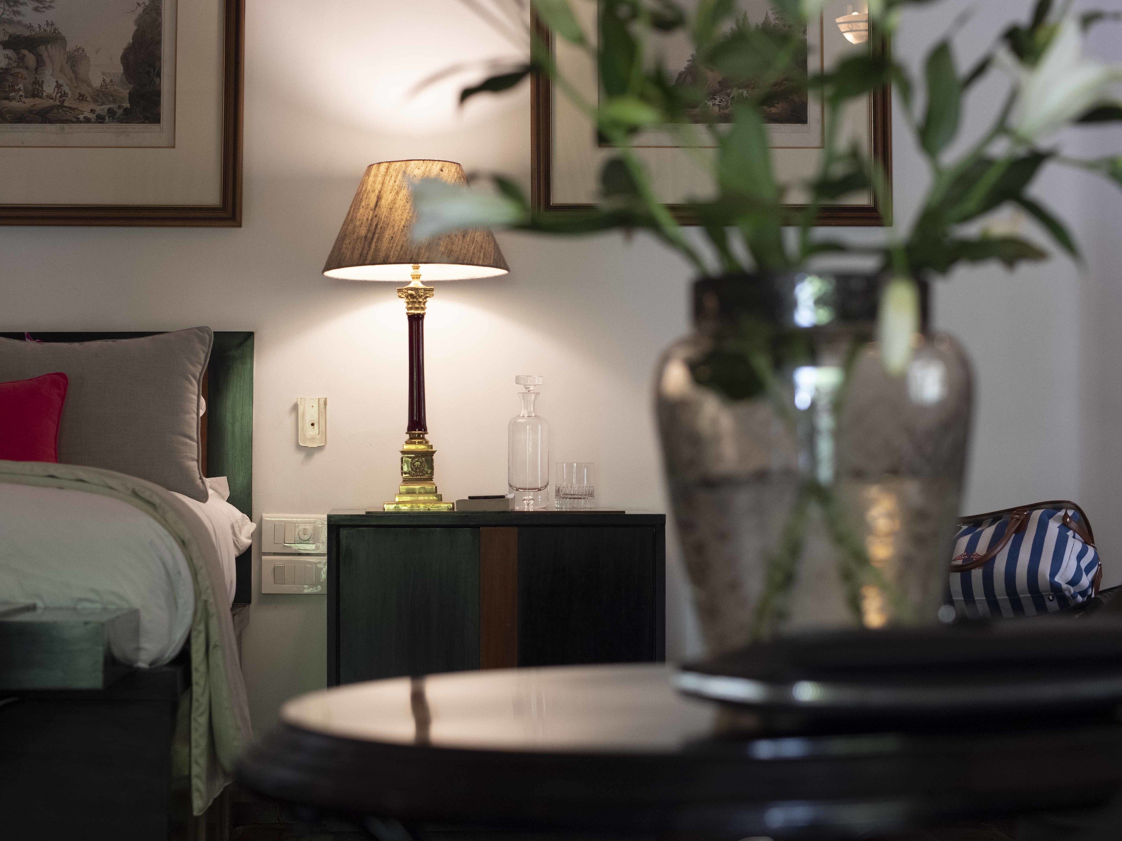 Una habitación de hotel con una cama, una mesita de noche con una lámpara, un jarrón con plantas y cuadros en la pared.