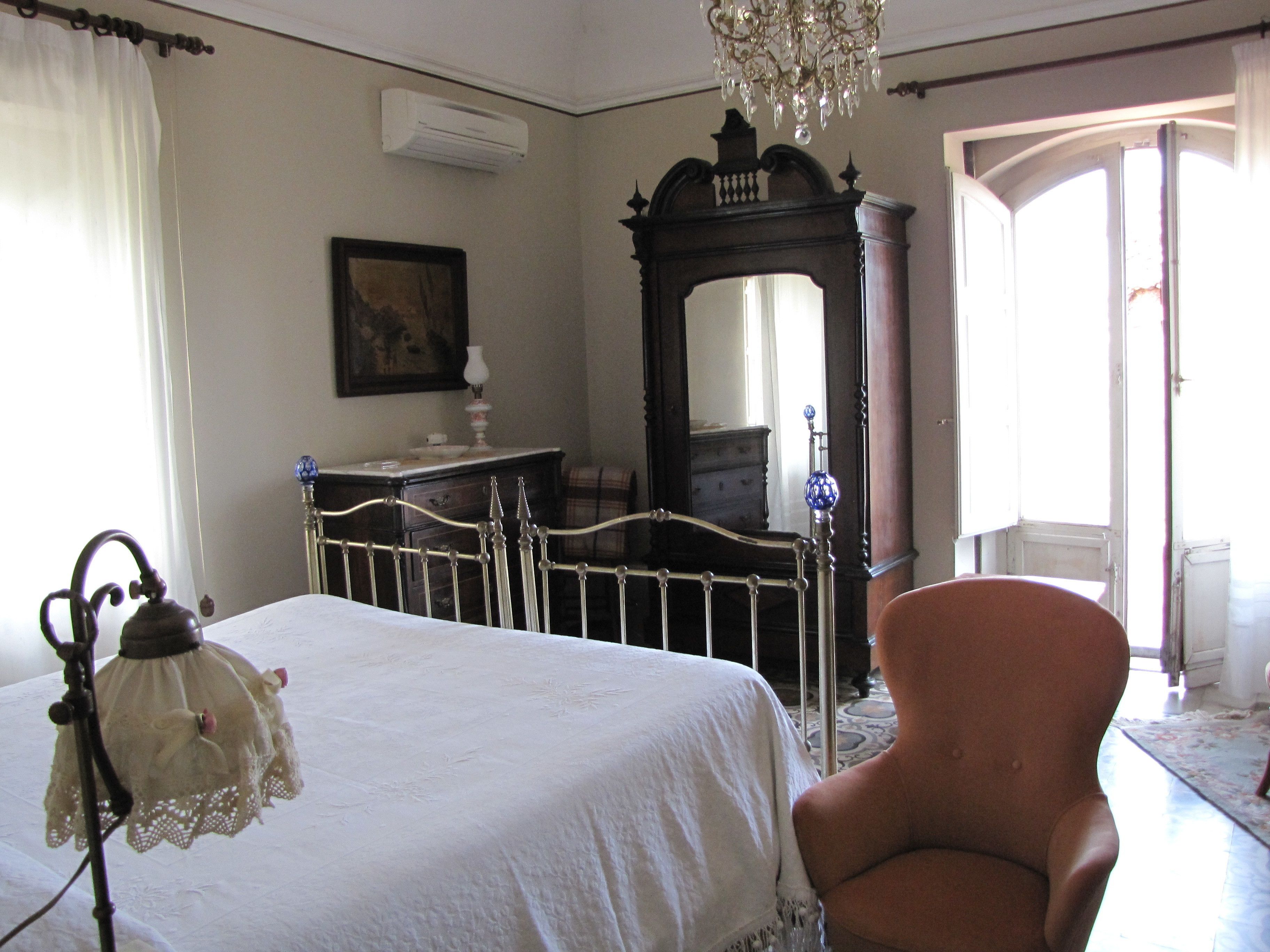 Un dormitorio con cama, una silla, tocador, espejo y candelabro.