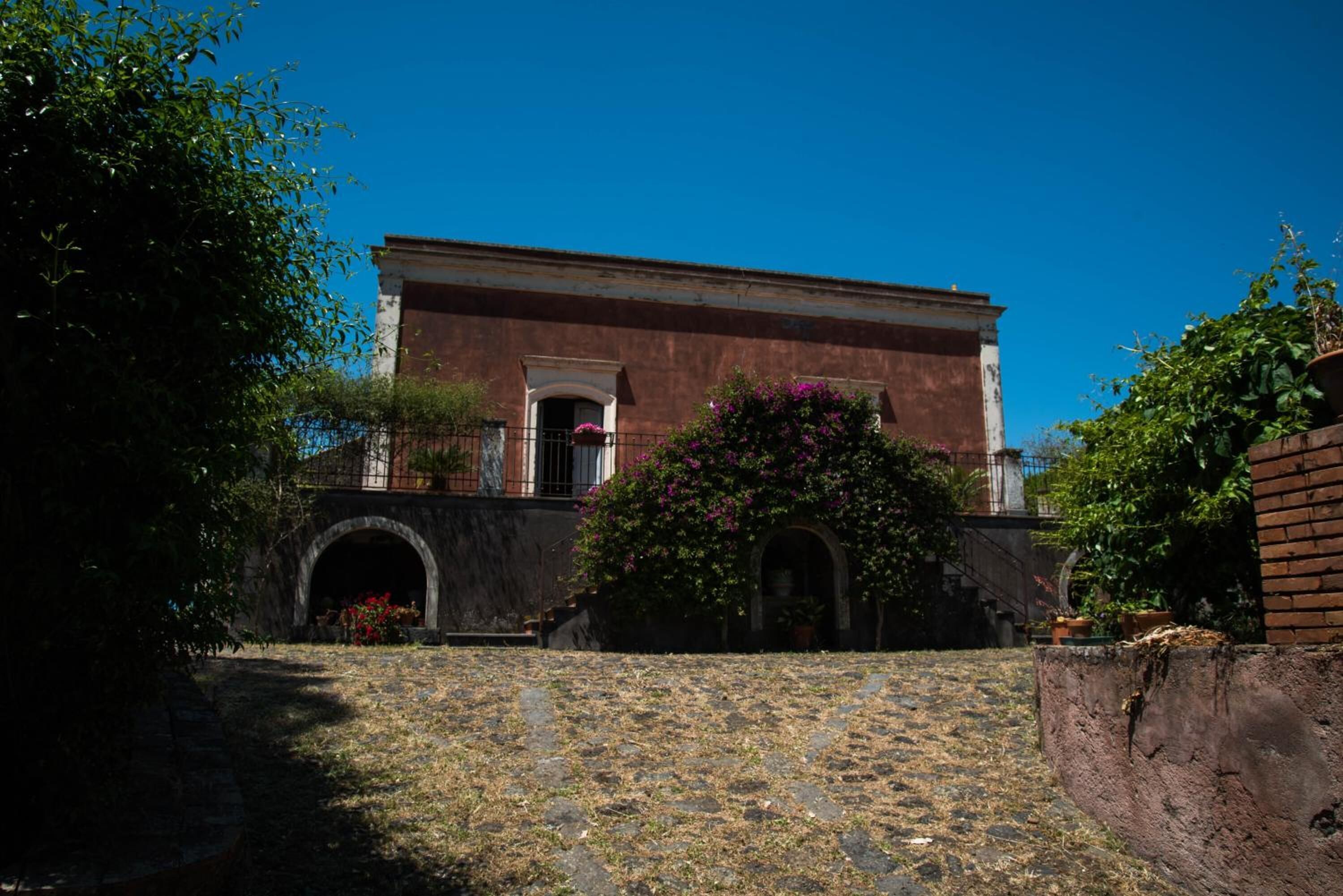 A historic building with a central archway, a small balcony, and a garden area with flowering plants.