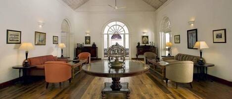 A spacious room with a high ceiling, wooden flooring, and a central round table surrounded by chairs.