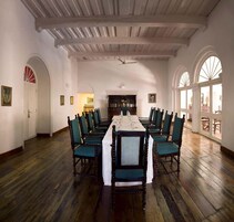 A dining room with a long table set for a meal, surrounded by chairs, with a bookshelf and arched windows in the background.