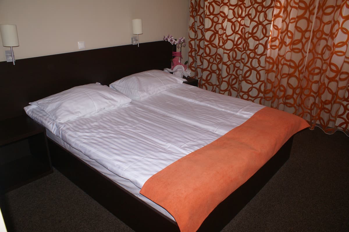 A neatly made bed with white linens and an orange throw blanket, a nightstand with a lamp, and a patterned curtain.