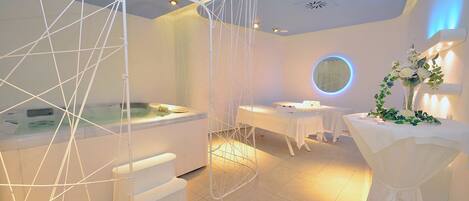 A modern spa room with a hot tub, a white chair, and a table with a floral arrangement.