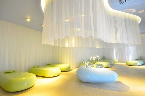 Un intérieur moderne avec des coins salons originaux aux couleurs verte et bleue, une table blanche centrale agrémentée de bougies et un plafond décoré de panneaux de tissu blanc suspendus.
