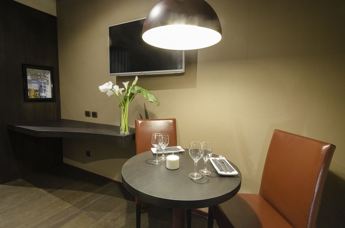 A dining area with a round table set for two, a vase with white flowers, a flat-screen TV mounted on the wall, and a modern pendant light.