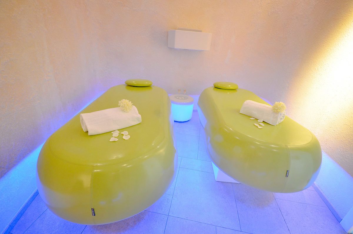 Two green massage chairs with towels and flowers, a white shelf with a box, and a blue-lit floor.