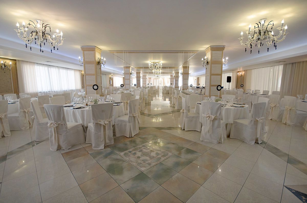 A spacious banquet hall with multiple round tables, white chairs, and chandeliers.