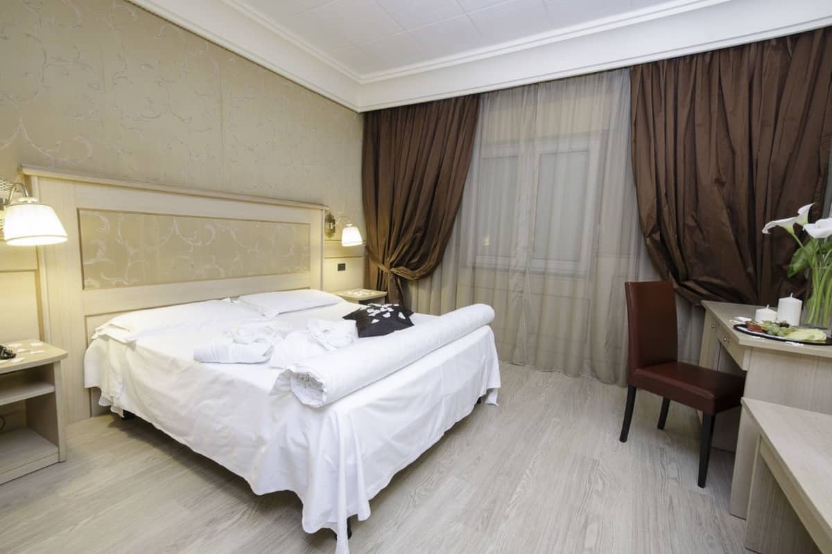 A hotel room with a large bed, two bedside tables with lamps, a desk with a chair, and a window with curtains.