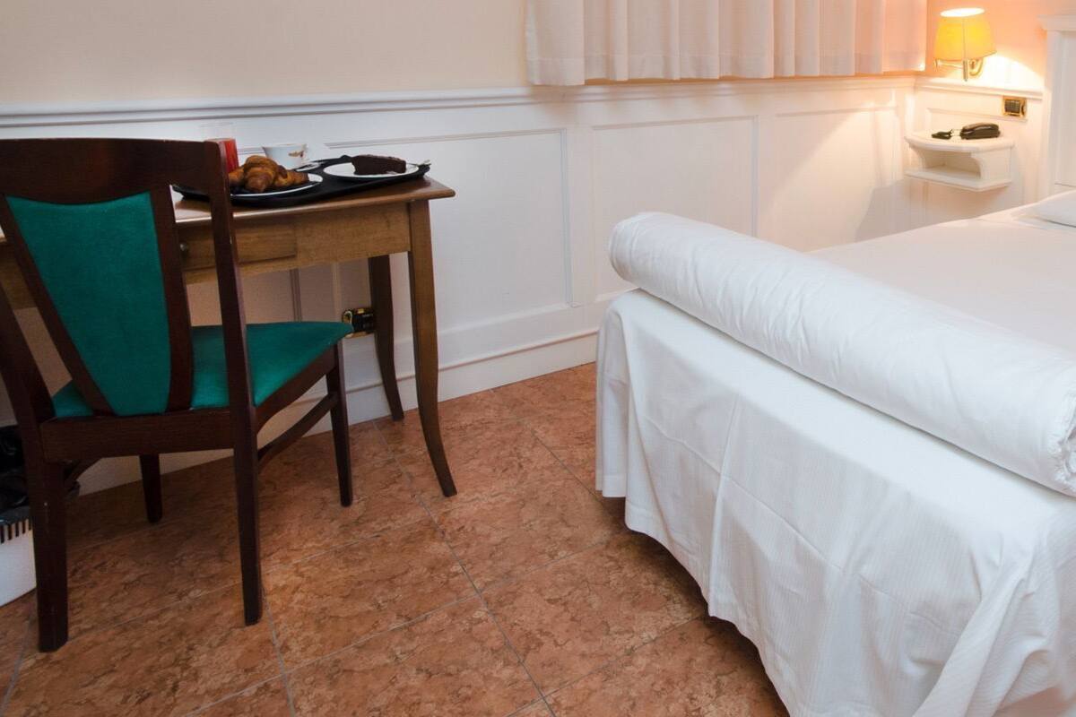 A single bed with white linens, a bedside table with a lamp, and a chair with a green cushion.
