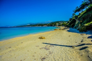 Une plage de sable fin bordée d’une eau d’un bleu limpide, un promontoire rocheux et une colline couverte de végétation.