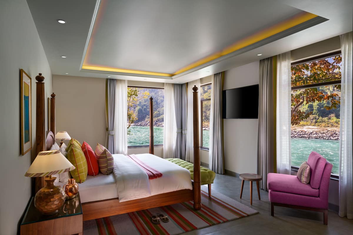 A bedroom with a large bed, a pink armchair, a small table, and a view of a body of water through large windows.