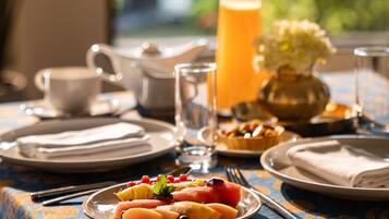 A breakfast table with plates of fruit, a glass of orange juice, and a pitcher of juice, set against a window with a view of greenery.