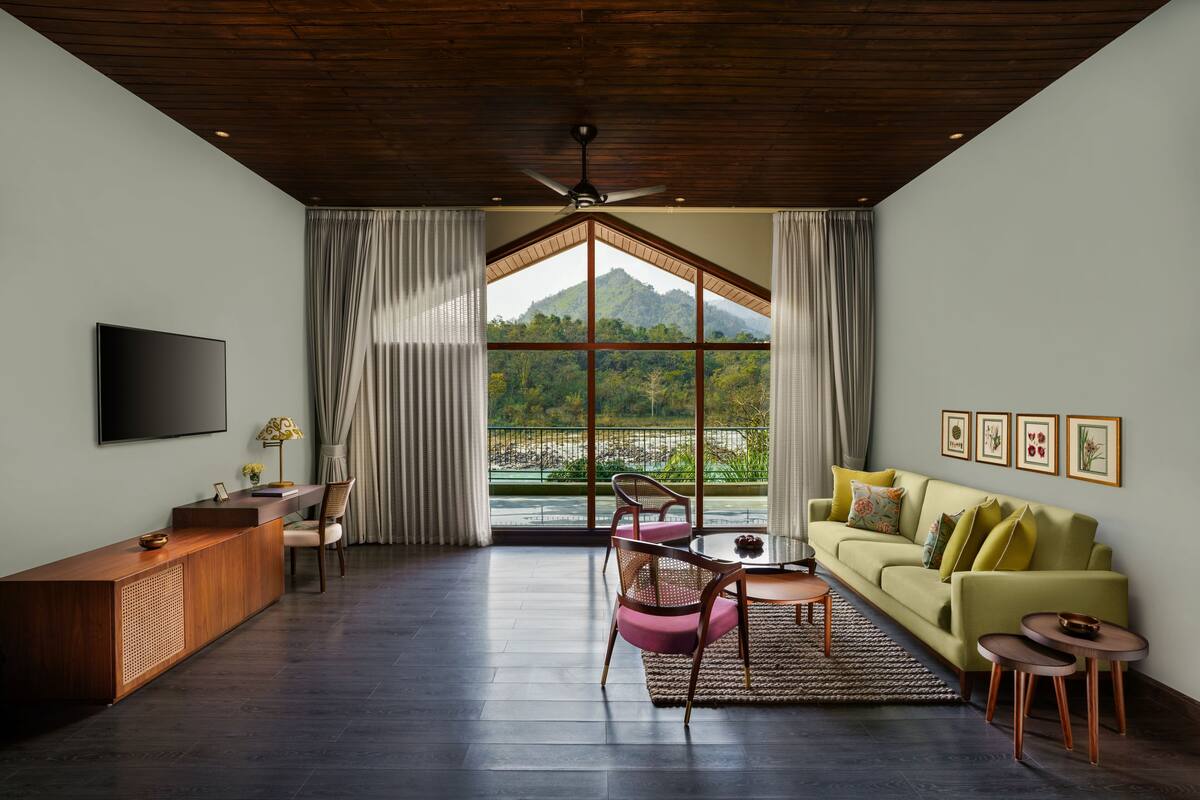 A modern living room with a wooden ceiling, a flat-screen TV, a sofa, a dining table, and a view of a mountain through large windows.