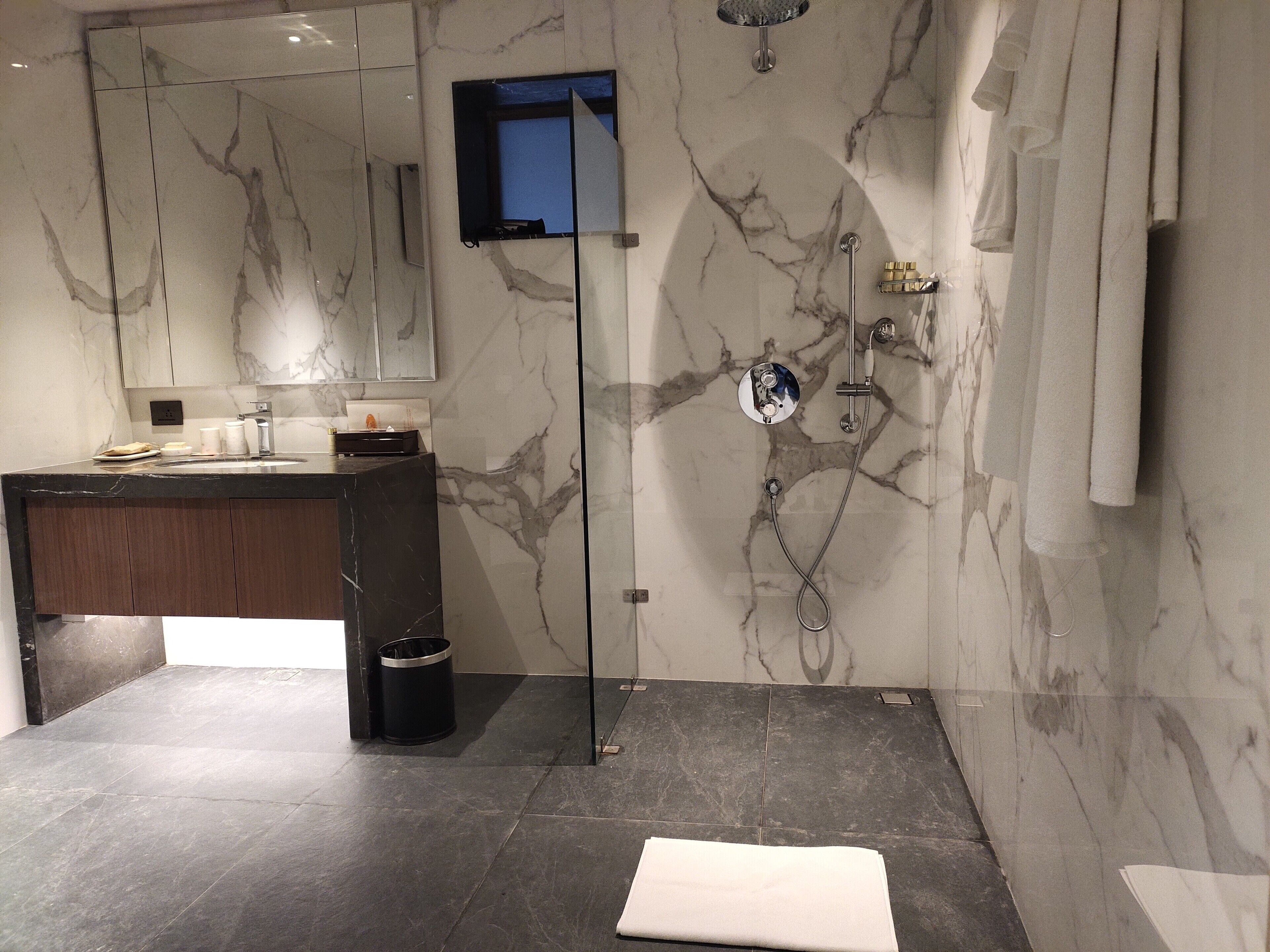 A modern bathroom with a marble wall, a glass shower enclosure, a dark wood vanity, and a white towel on the floor.