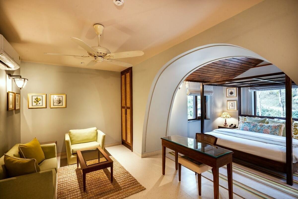 A room with a bed, a desk, two chairs, a ceiling fan, and a large arched window.