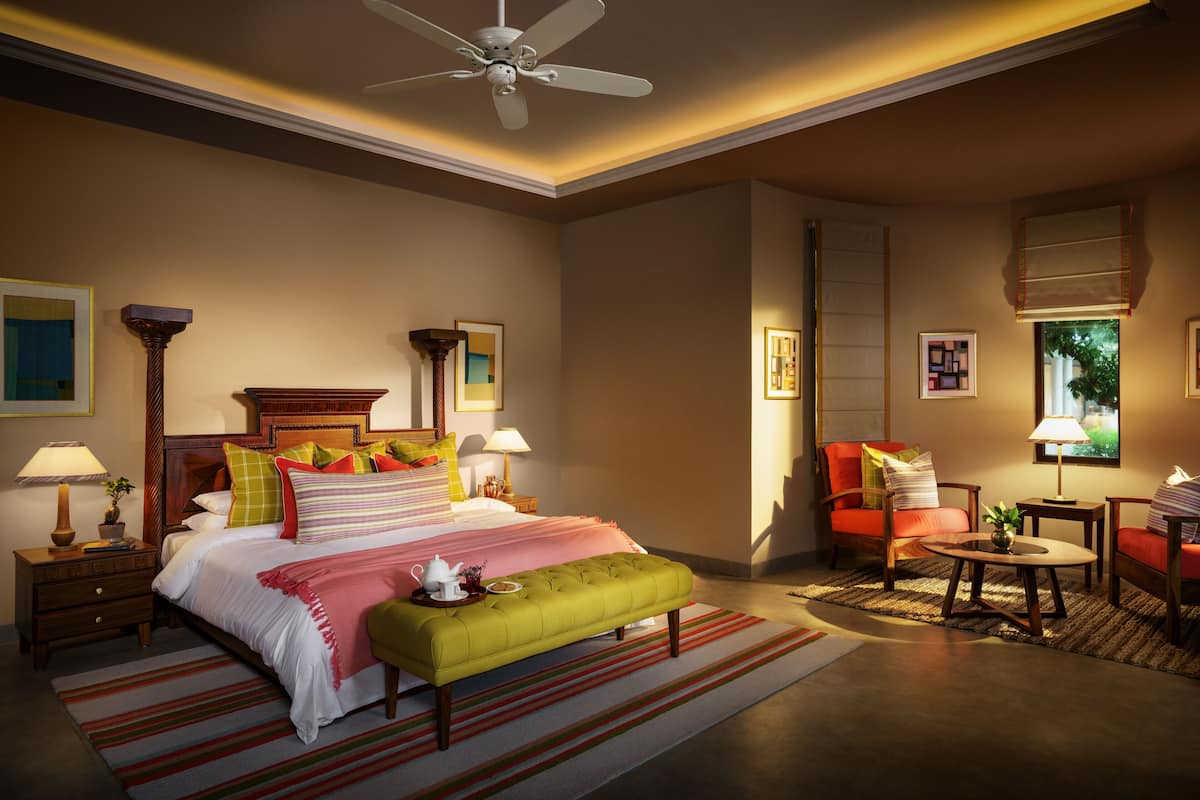 A well-lit bedroom with a bed, bedside tables, a bench, a chair, a small table, and a ceiling fan.