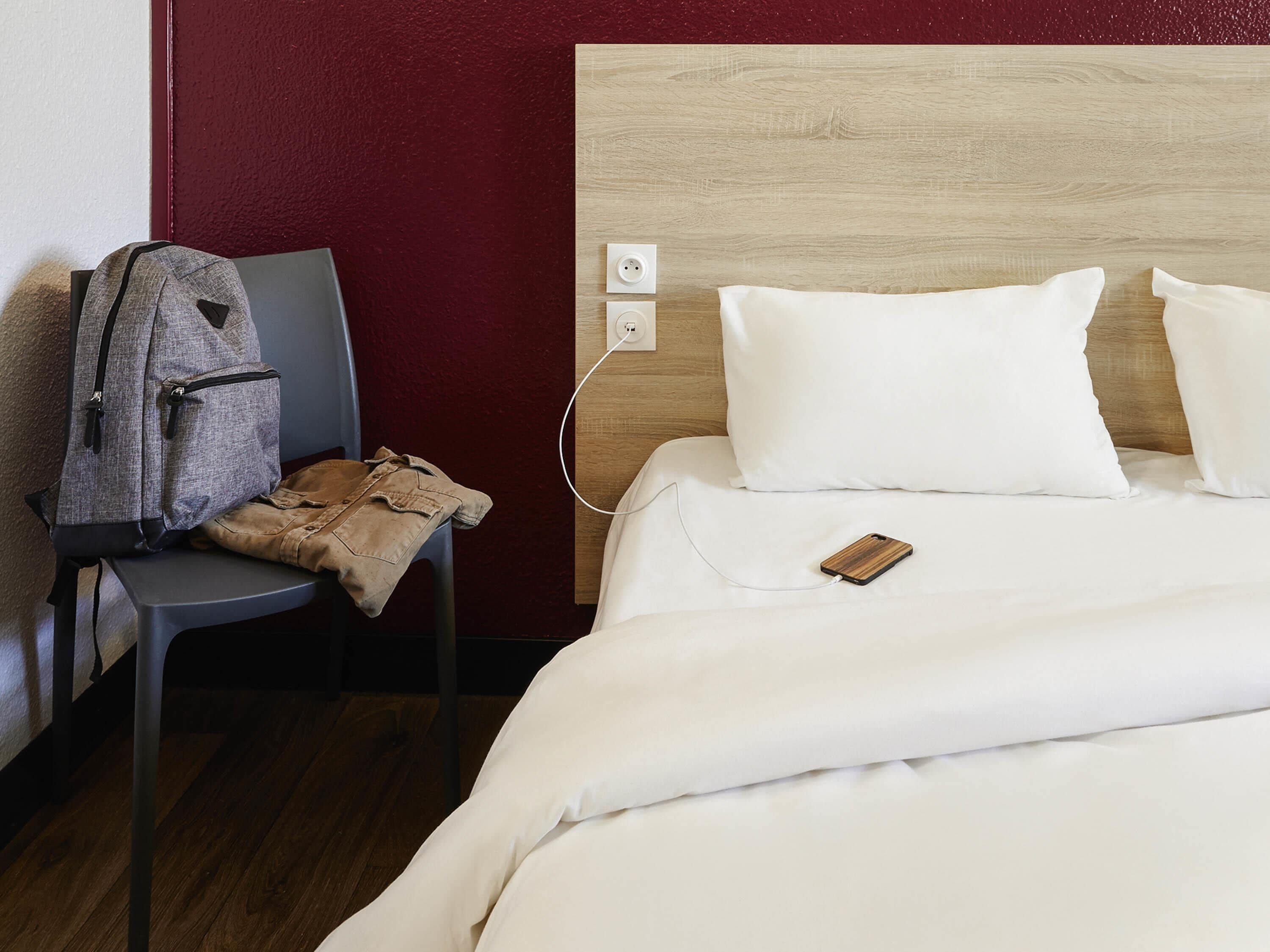 A hotel room with a wooden headboard, white bedding, a gray chair, a backpack, and a smartphone on the bed.
