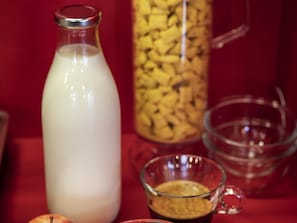 A bottle of milk, a glass mug with a beverage, and a glass pitcher on a red surface.