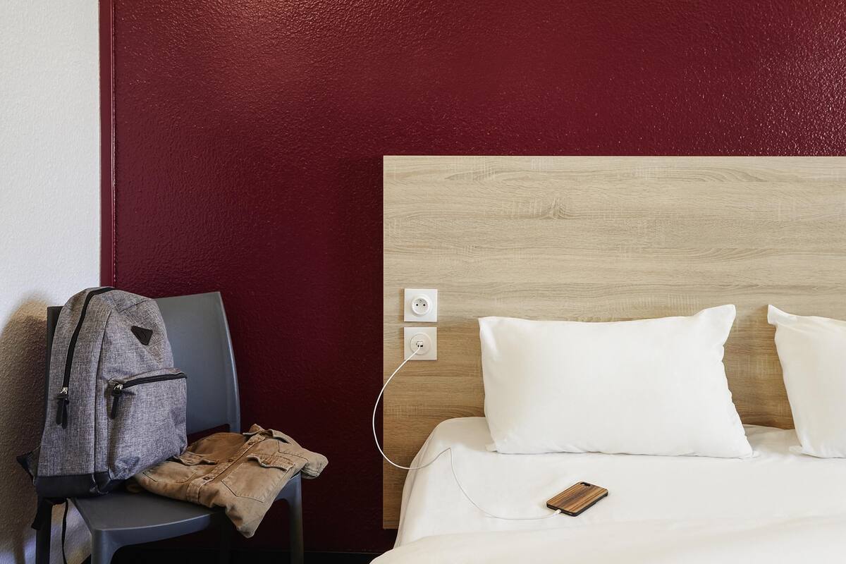 Ein Hotelzimmer mit Bett, Sessel, Rucksack und einer Steckdose.
