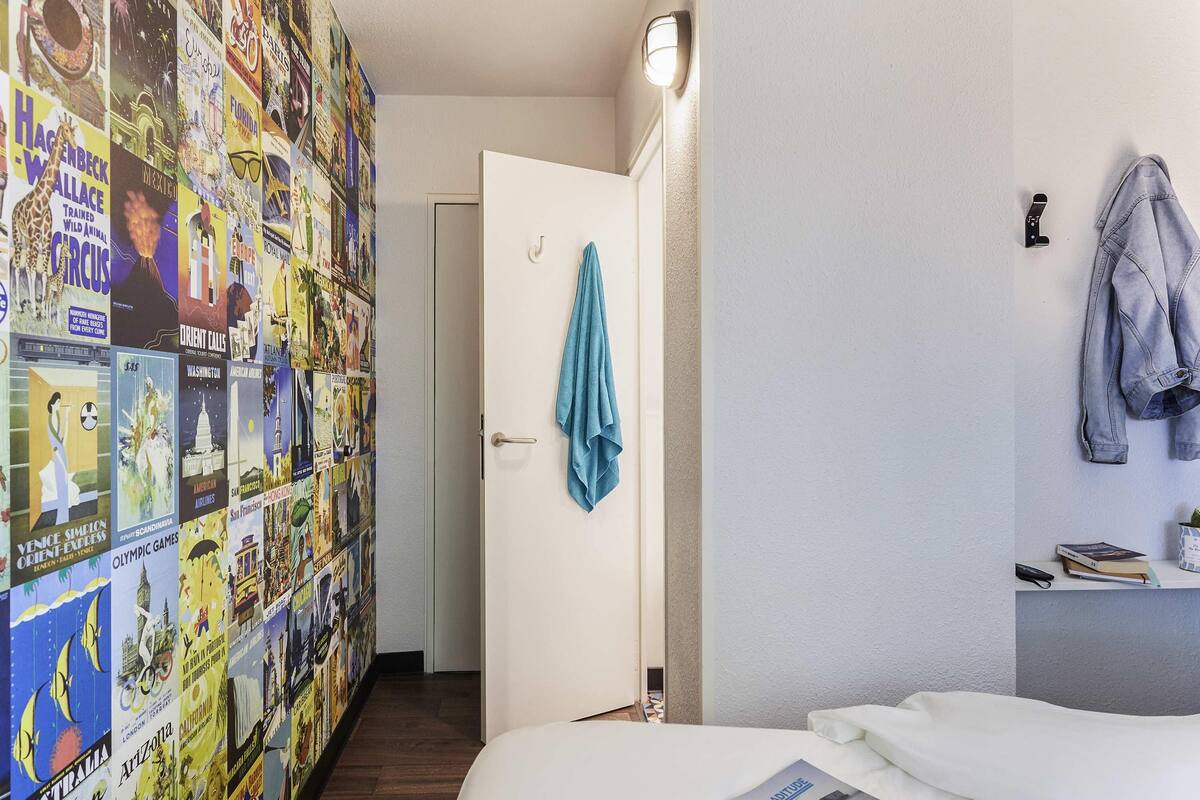 Ein Hotelzimmer mit einer mit Reiseplakaten verzierten Wand, einem Bett mit einer Zeitschrift, einer Tür mit einem aufgehängten Handtuch und einer Leuchte.