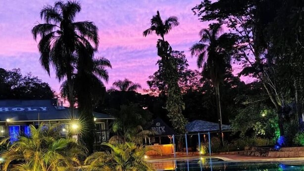 Una piscina con borde de piedra, rodeada de palmeras y un edificio con la palabra 'HOLY' escrita.