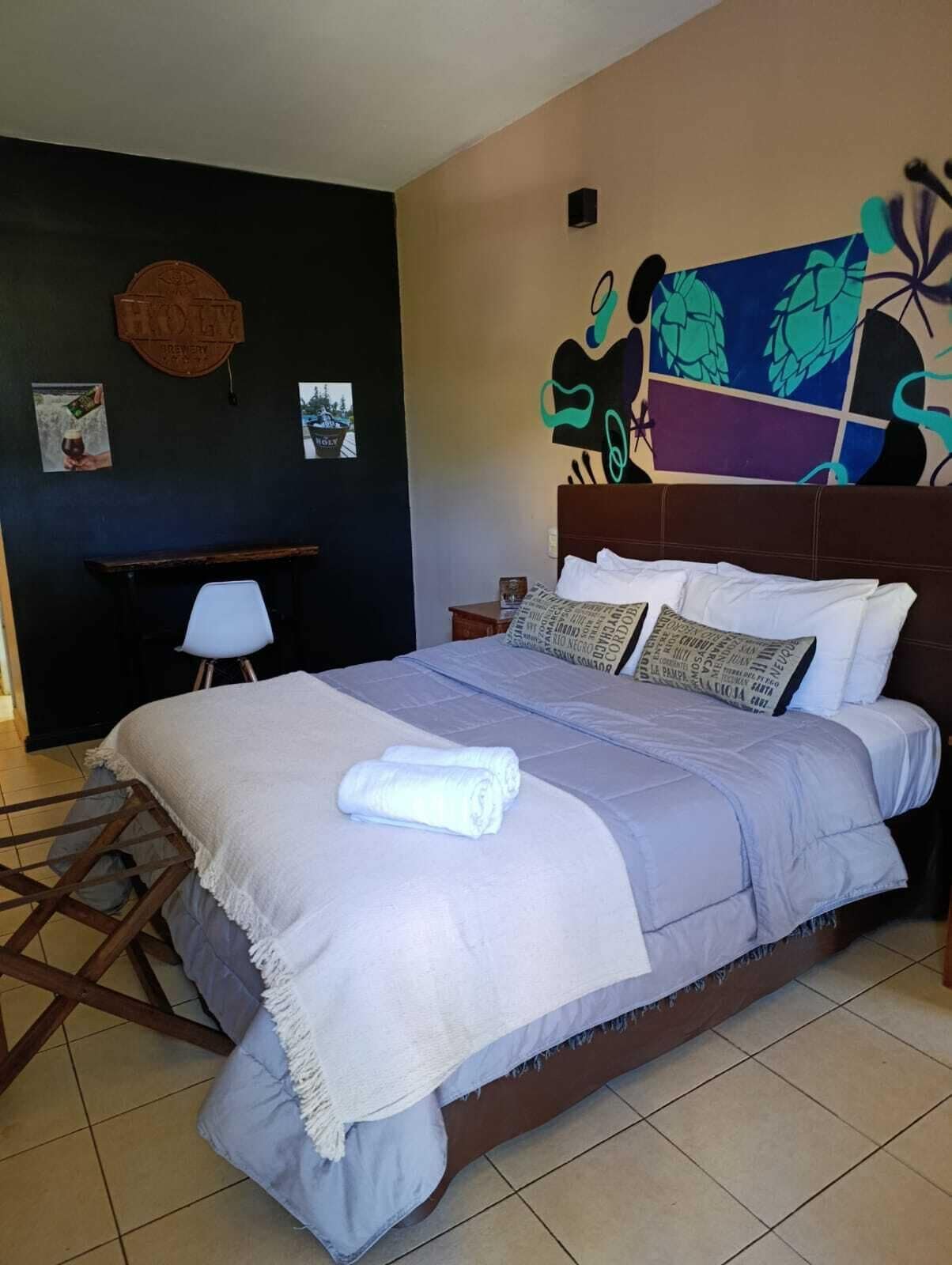 A neatly made bed with a white duvet and rolled towels, a wooden headboard, and a wall with a colorful abstract painting.