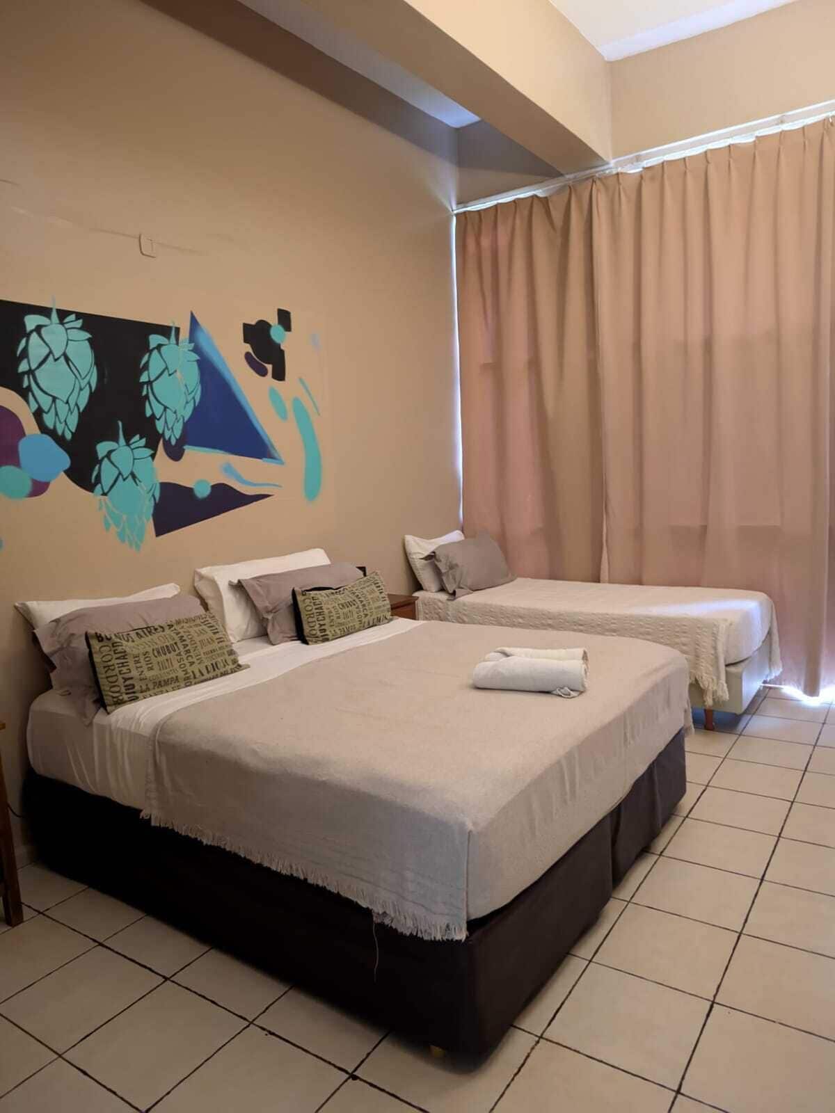 A hotel room with two beds, a large abstract wall mural, and a window with beige curtains.