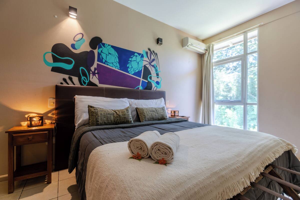 A neatly made bed with rolled towels, a decorative wall art piece, and a window with a view of trees.