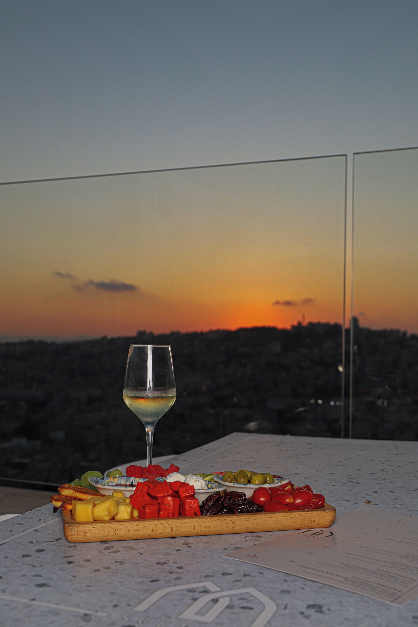 Una mesa con una copa de vino blanco y una tabla de madera con frutas y verduras variadas.