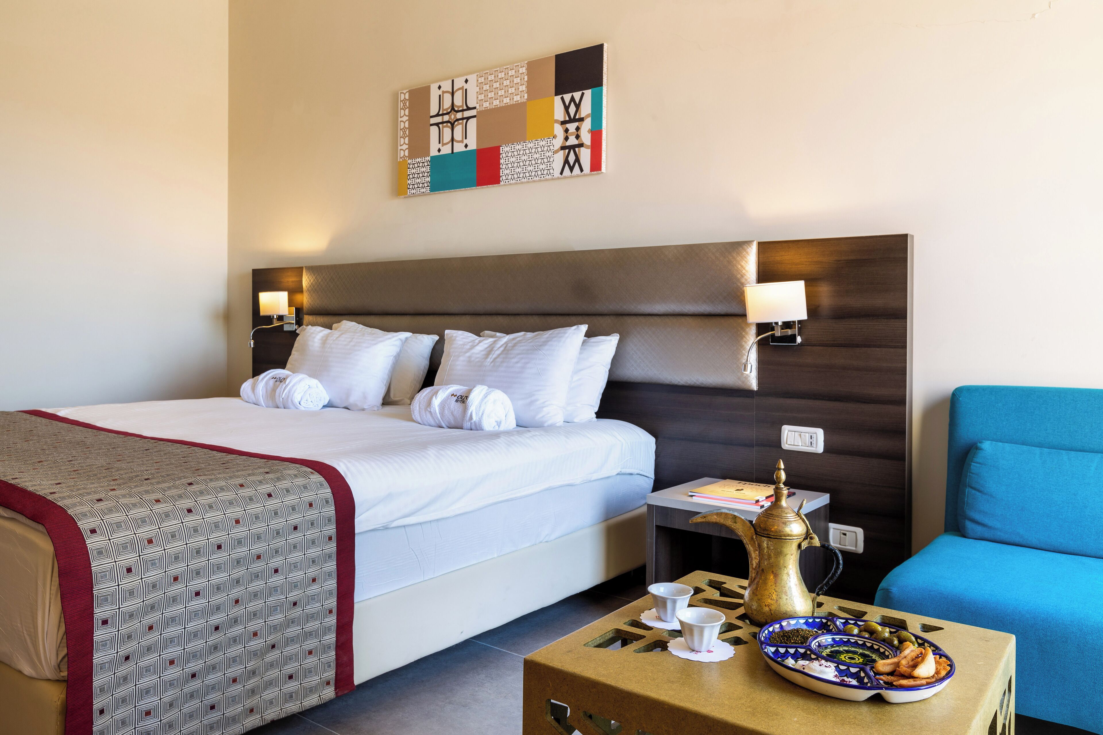 Habitación de hotel con cama, lámparas de noche, un sillón azul, una mesita con cafetera y tazas, y un elemento decorativo en la pared.