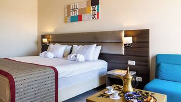 Een hotelkamer met een bed, bedlampen, een blauwe fauteuil, een klein tafeltje met een koffiezetapparaat en kopjes, en een decoratief wandobject.