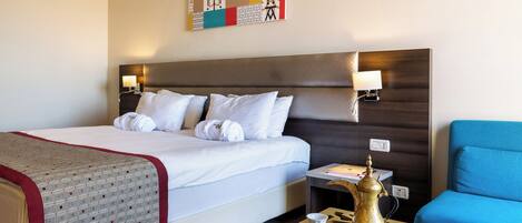 Habitación de hotel con cama, lámparas de noche, un sillón azul, una mesita con cafetera y tazas, y un elemento decorativo en la pared.