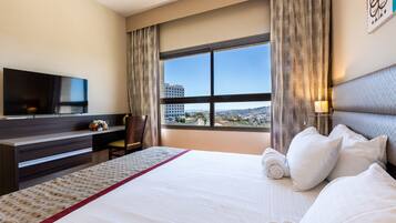 A hotel room with a large bed, a desk with a flat-screen TV, and a view of the city through the window.