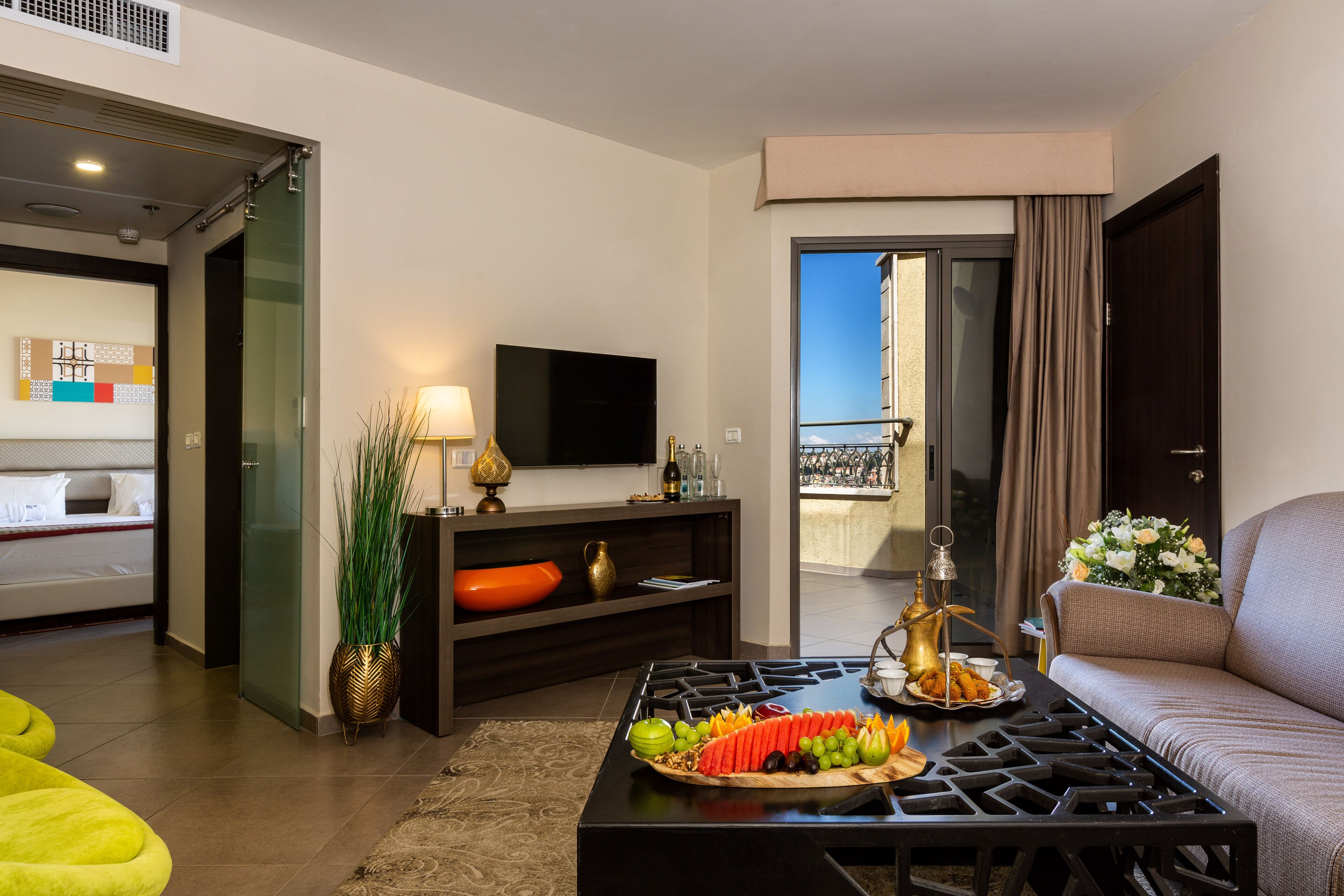 A modern living room with a flat-screen TV, a dining table with fruit, a sofa, and a balcony view.