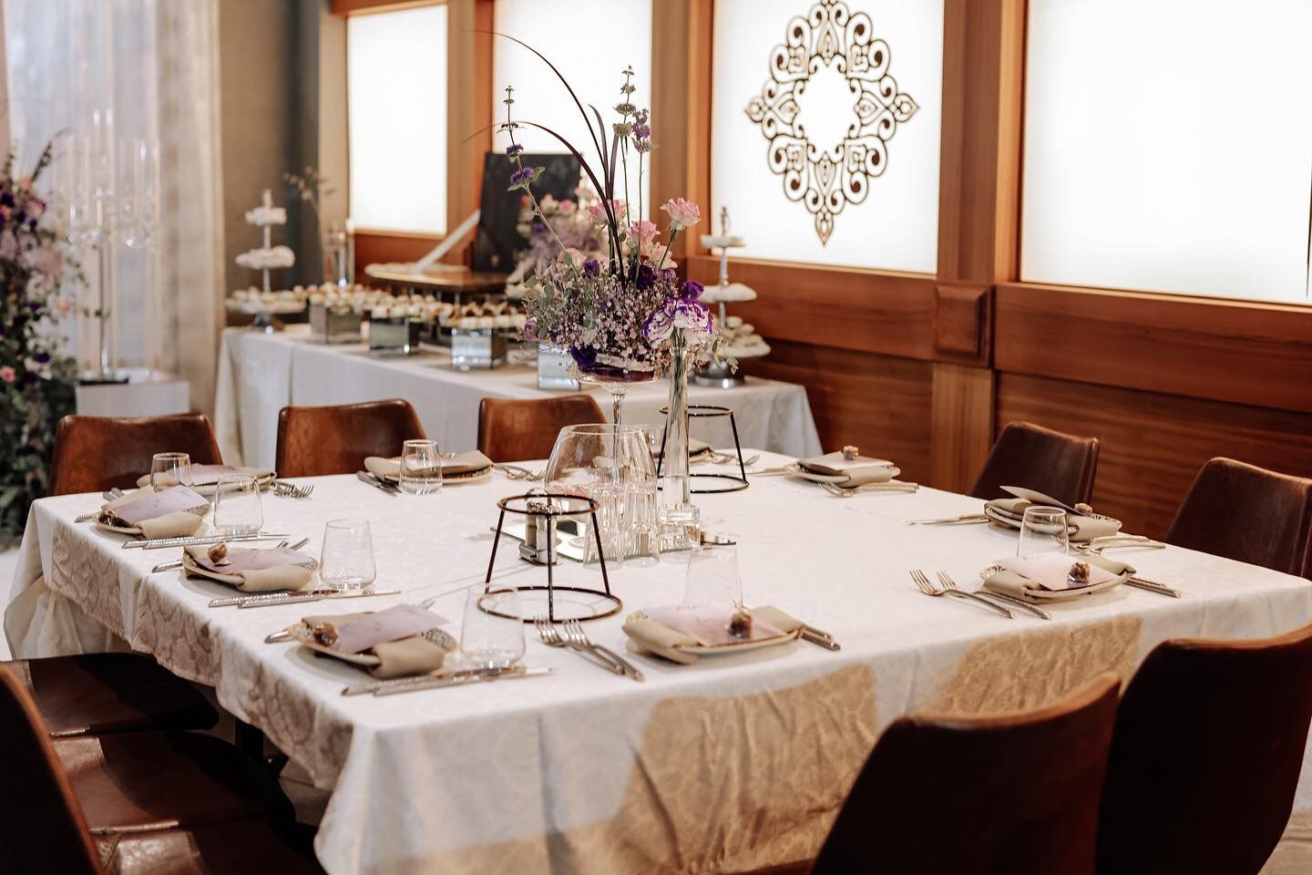 A formal dining room with a long table set for a meal, featuring white tablecloths, neatly arranged plates, glasses, and cutlery.