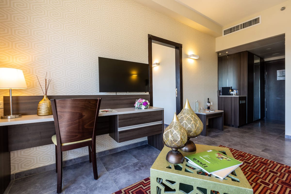 Ein Hotelzimmer mit einem Holztisch, einem Sessel, einem Fernseher, einer dekorativen Lampe und einem kleinen Tisch mit einer Vase und einem Buch.