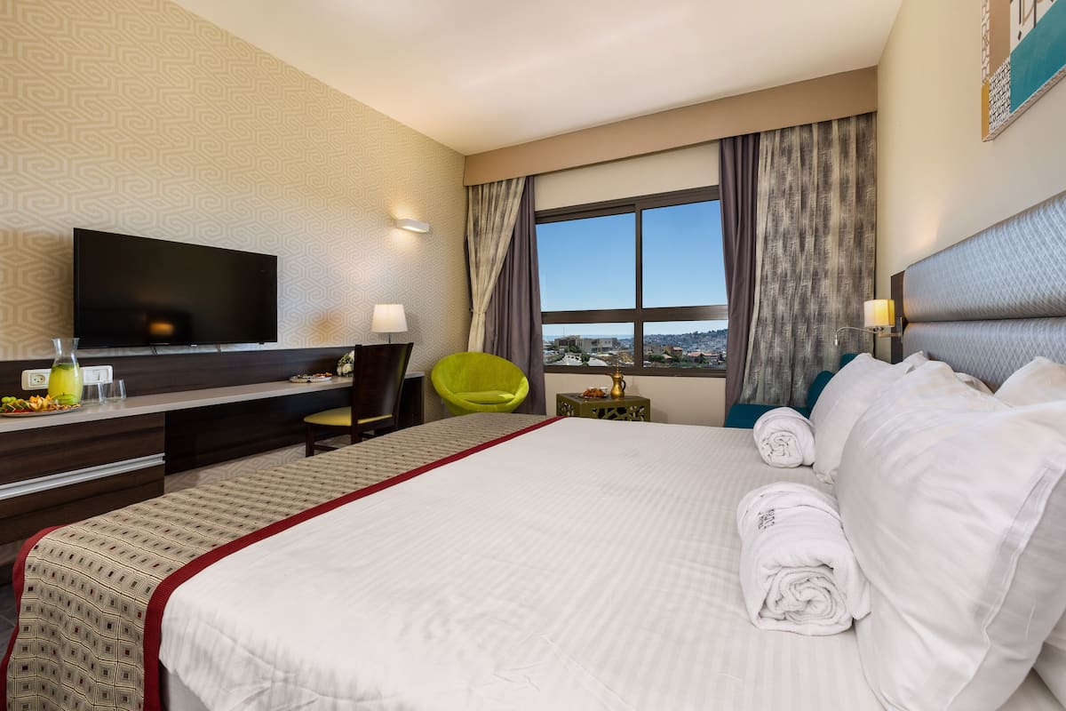 Ein Hotelzimmer mit einem großen Bett, einem Flachbildfernseher, einem Schreibtisch mit Stuhl und Blick auf die Stadt durch das Fenster.