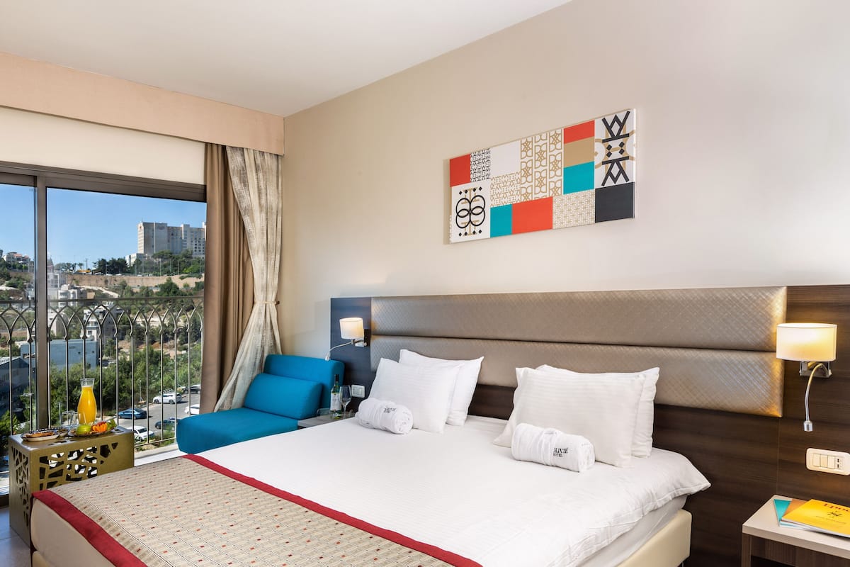 Ein Hotelzimmer mit einem großen Bett, einem blauen Sessel, Blick auf die Stadt und einem modernen, abstrakten Kunstwerk an der Wand.