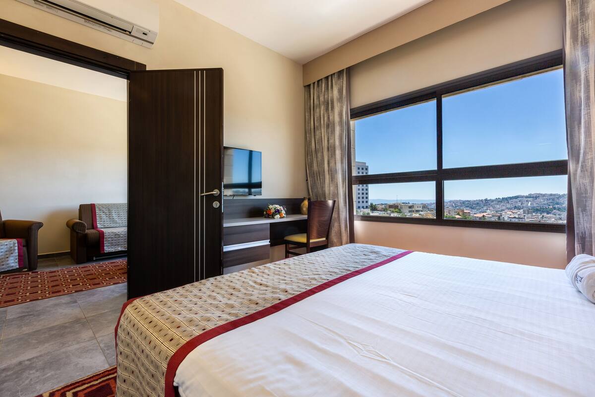 Ein Hotelzimmer mit Bett, Schreibtisch, Stuhl und einem großen Fenster mit Blick auf die Stadt.